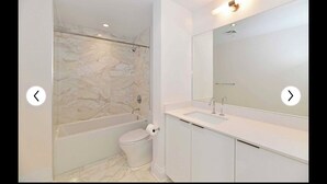Une salle de bain moderne avec un mur de douche en marbre, des toilettes blanches et un meuble-lavabo doté d’un grand miroir.