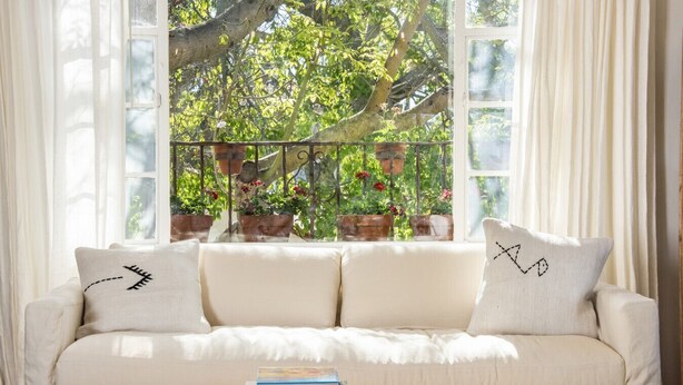 A white sofa with two pillows, a round woven coffee table, and a view of a garden through a window with curtains.