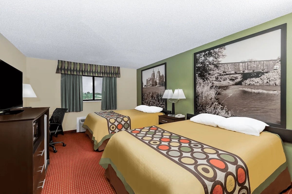 A hotel room with two beds, a desk with a TV, a window with curtains, and a large picture of a bridge and river.