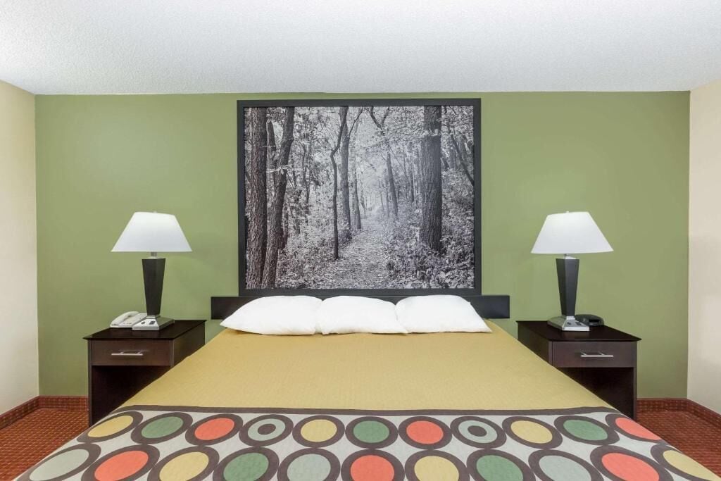 A hotel room with a bed, two bedside tables with lamps, a telephone, and a large forest-themed artwork on the wall.