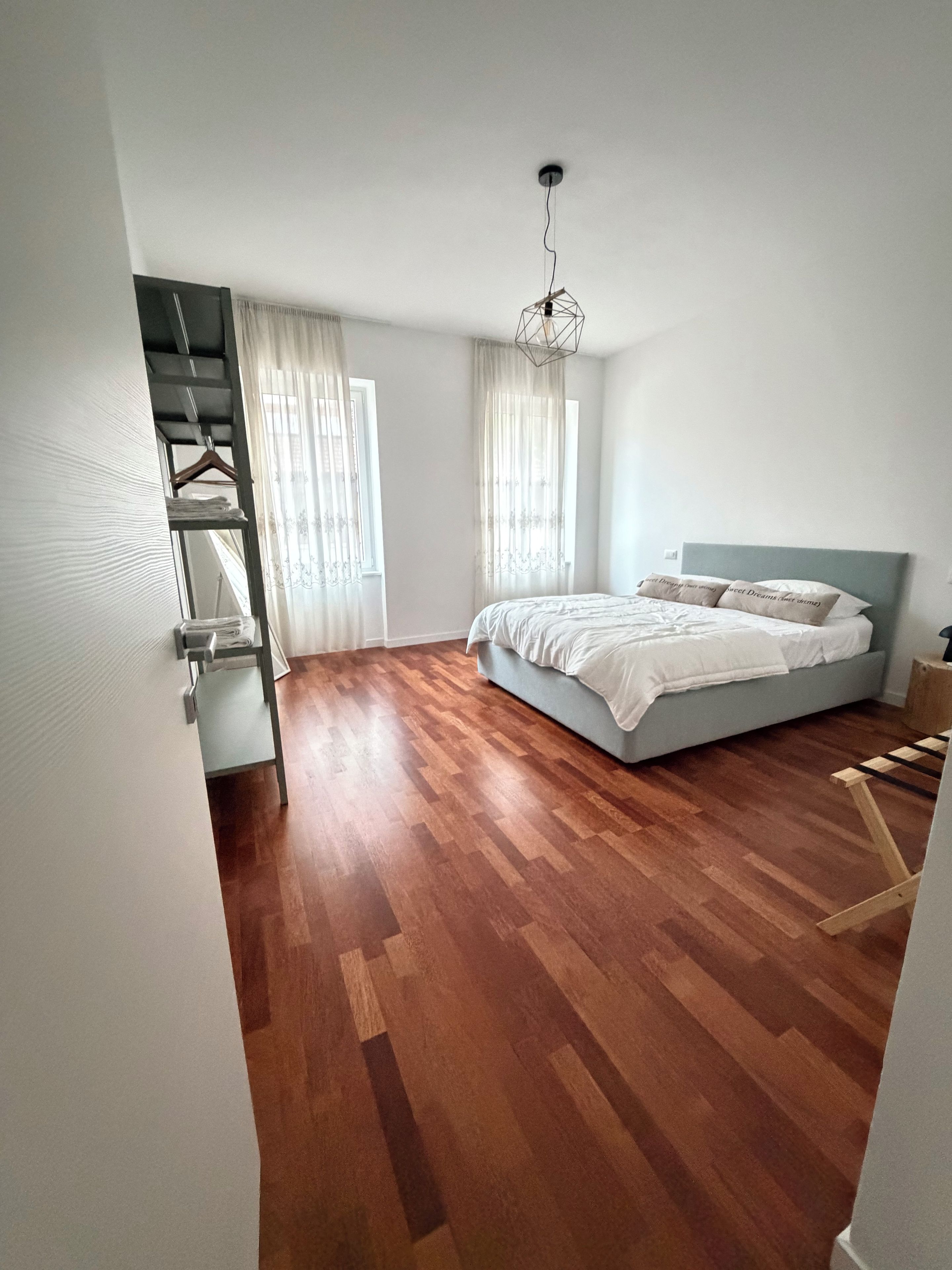A bedroom with a bed, wooden flooring, a hanging light fixture, and a window with curtains.
