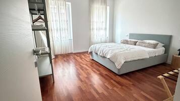 A bedroom with a bed, wooden flooring, a hanging light fixture, and a window with curtains.