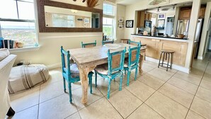 A dining area with a wooden table and turquoise chairs, a kitchen with modern appliances, and a view through a window.