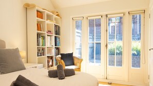 Une chambre avec un grand lit, une bibliothèque, un fauteuil en osier et une vue sur l’extérieur, visible à travers la porte.