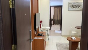A modern hotel room with a wooden desk, a flat-screen TV, a small table, and a chair. There is a fire extinguisher on the wall and a painting above the door.