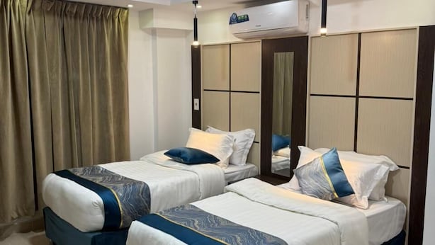 A hotel room with two beds, beige tiled flooring, a large window with curtains, and an air conditioning unit on the wall.