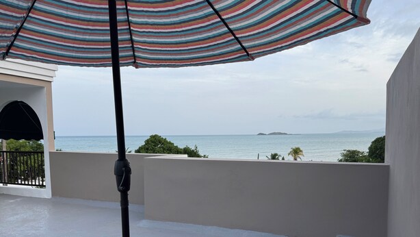 A balcony with a view of the sea, two red chairs, and a striped umbrella.