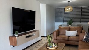 A modern living room with a flat-screen TV, a brown leather sofa, a wooden coffee table, and a wicker pendant light.