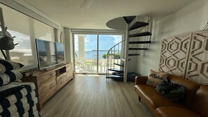 Un salon moderne avec un téléviseur, des meubles en bois, un canapé en cuir marron et un escalier en colimaçon menant à un balcon avec vue sur la mer.