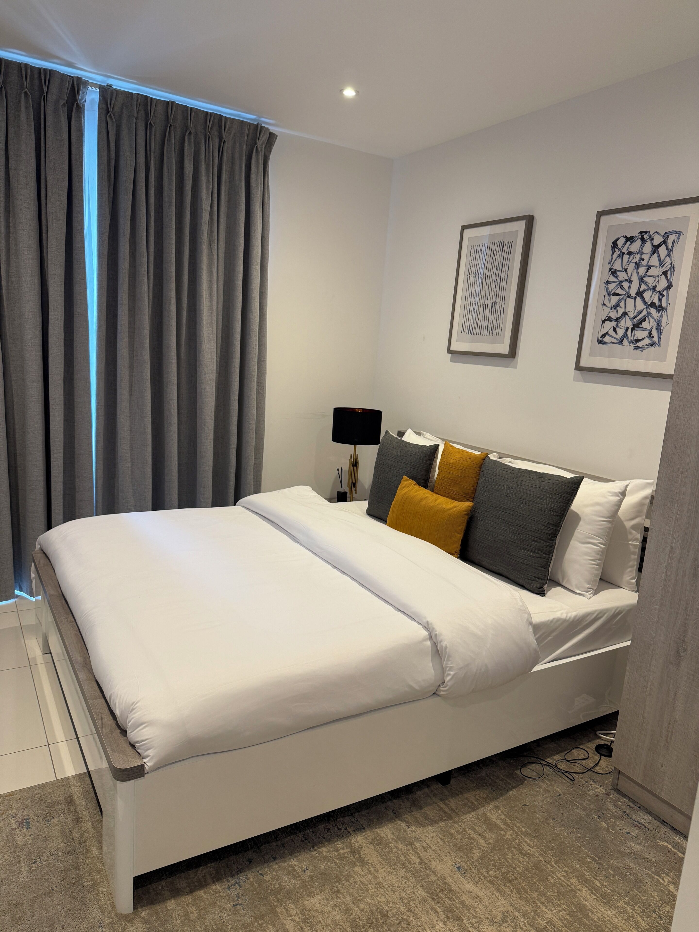 A modern bedroom with a white bed, grey curtains, and two abstract art pieces on the wall.