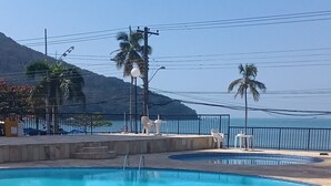 A clear blue swimming pool with a tiled deck, surrounded by a fence and palm trees. - Seafront Apartment with Pool in Martim de Sá (Caraguatatuba)