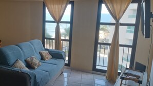 A living room with a blue sofa, a wooden dining table, and two windows with curtains. - Seafront Apartment with Pool in Martim de Sá (Caraguatatuba)