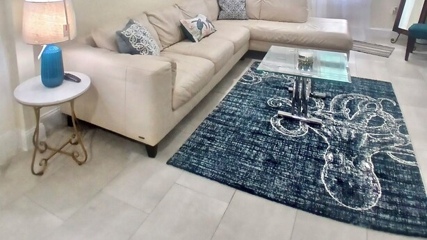 En stue med en beige sofa, et sofabord, en blå måtte og et billede på væggen.