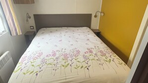 A compact bedroom with a bed, a small bedside table, a wall-mounted shelf, and a window with curtains. - Mobile home rental 8 berths (Parentis-en-Born)