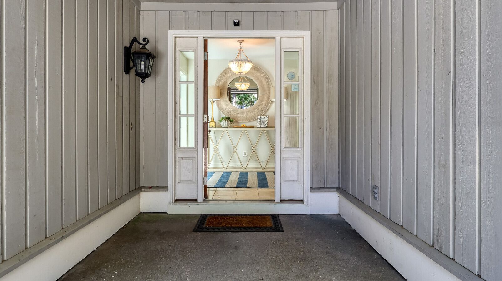 A hallway with a door leading to a room with a chandelier, a mirror, and a small shelf with decorative items.