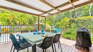 A covered outdoor patio with a table and chairs, surrounded by lush greenery.