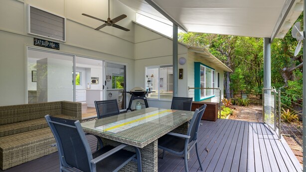 Un espace extérieur abrité, aménagé pour les repas, avec une table et des chaises, un ventilateur de plafond et une vue sur la maison et le jardin.
