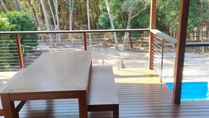 Una terraza de madera con una mesa de picnic, una piscina y un área cercada con árboles al fondo.