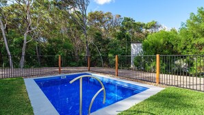 Una piscina rectangular con revestimiento azul, rodeada por una valla de metal negro, y con árboles y follaje de fondo.
