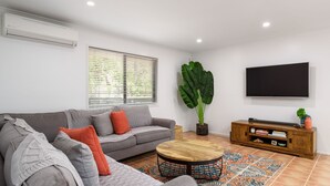 Ein Wohnzimmer mit grauem Sofa, orangefarbenen Kissen, einem Holztisch, einer großen Grünpflanze, einem Fernseher und einer Holzkommode mit Büchern.
