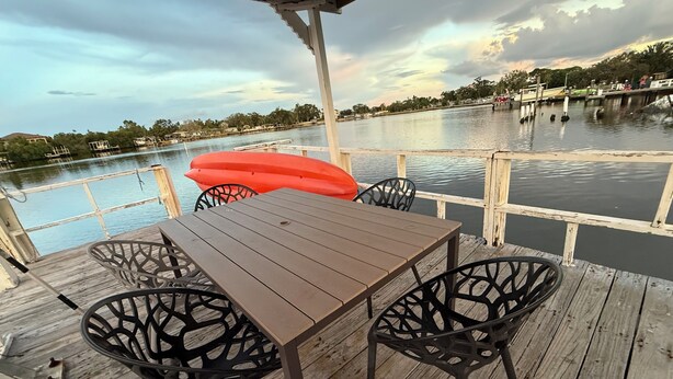 Una terraza de madera con una mesa y sillas, con vista a un cuerpo de agua tranquilo que incluye un muelle al fondo.
