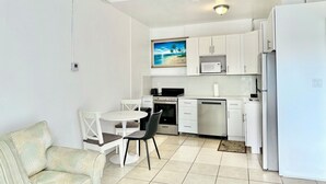 Una cocina compacta con armarios blancos, microondas, estufa y refrigerador. Hay una mesa de comedor con sillas y un sofá en el área de estar contigua.