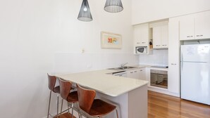 Eine Küche mit einer zentralen Kochinsel, weißen Schränken, einer Mikrowelle, einem Backofen und einem Kühlschrank. Um die Insel sind drei Holzstühle gestellt.