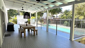 Ein überdachter Außenbereich mit Holzterrasse, einem Tisch mit Bänken, einer hängenden Leuchte und Blick auf einen Pool.