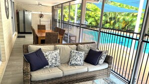 A wicker sofa with cushions, a dining table, chairs, and a view of a pool.