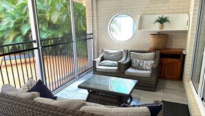 A spacious balcony with wicker furniture, a glass coffee table, and a view of palm trees.