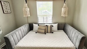 Un grand lit doté d’une tête de lit et d’un pied de lit recouverts d’un tissu gris, d’une tête de lit en bois et de deux décorations murales en macramé.