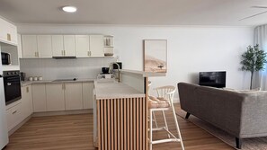 A modern kitchen with white cabinets, a wooden bar stool, a grey sofa, and a TV.