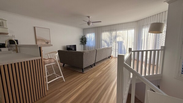 A modern living room with a sofa, a dining area with a white chair, a ceiling fan, and a staircase.