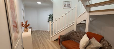 Una escalera que conduce a un nivel superior, un sofá de mimbre con cojines, un cuadro en la pared y una planta en la esquina.