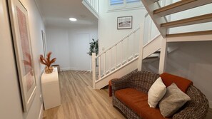 A staircase leading to an upper level, a wicker sofa with cushions, a framed picture on the wall, and a plant in the corner.