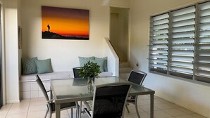Un coin repas avec une table en verre, des chaises, un canapé agrémenté de coussins à motifs, un tableau représentant un coucher de soleil et une fenêtre avec des stores.