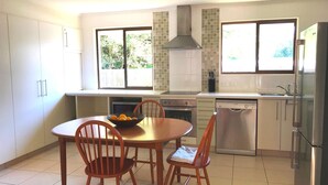 Une cuisine moderne avec des meubles blancs, une table à manger avec des chaises, un four encastré, un lave-vaisselle et une grande fenêtre donnant sur un paysage verdoyant.