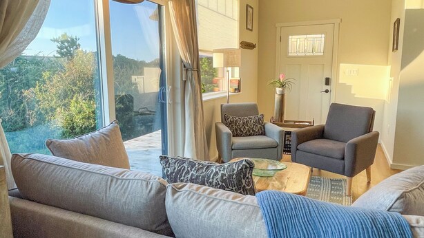 A spacious living area with a large sofa, two armchairs, and a coffee table. A sliding glass door leads to a balcony with a view of greenery.