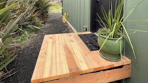 A wooden bench outside a building with a green door, surrounded by plants and a gravel path.