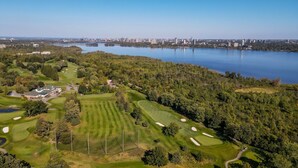 An aerial view of a golf course with a clubhouse, surrounded by dense trees and a city skyline in the background.