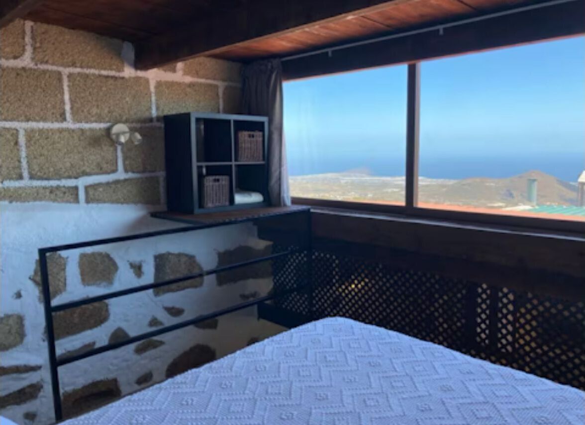 Een slaapkamer met een bed, een klein plankje en een raam met uitzicht op de oceaan en heuvels.