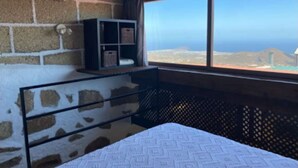 Un dormitorio con una cama, un estante pequeño y una ventana con vistas al océano y las colinas.