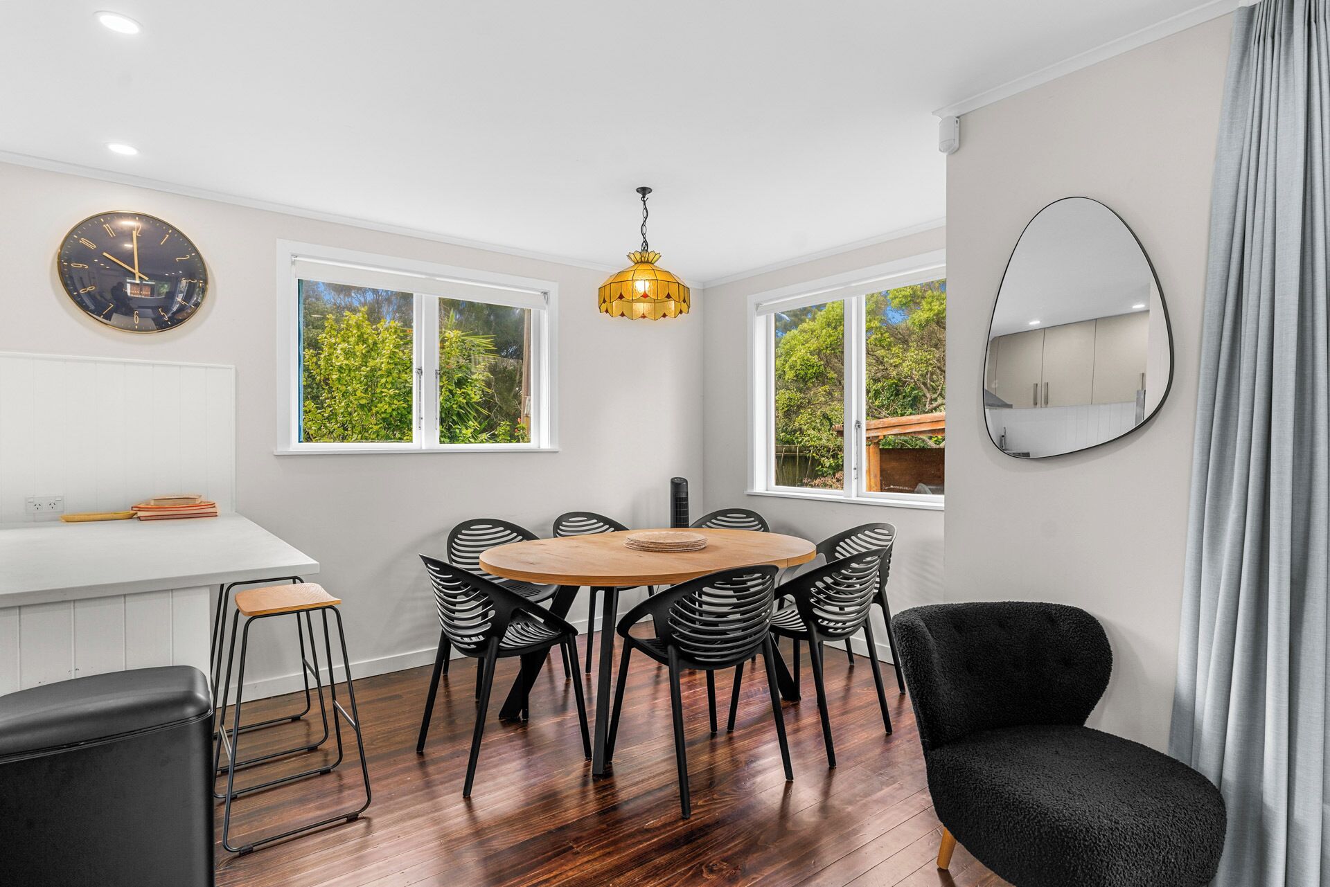 A modern kitchen with a round wooden table, black chairs, a large clock, and a hanging pendant light.