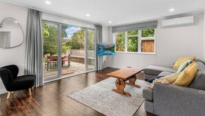 Ein modernes Wohnzimmer mit Holzboden, einer großen Schiebeglastür zum Garten, einem an der Wand montierten Flachbildfernseher und einem grauen Sofa mit gelben Kissen.