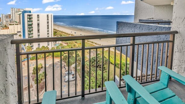 A balcony with a view of the beach and ocean, featuring a railing and two turquoise chairs.