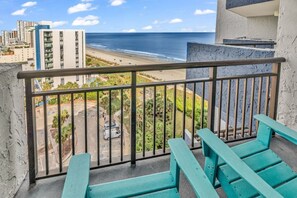 A balcony with a view of the beach and ocean, featuring a railing and two turquoise chairs.