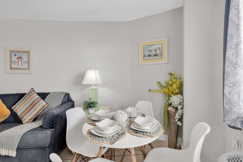 A modern living room with a grey sofa, a dining table set with white plates and glasses, a green lamp, and two framed pictures on the wall.