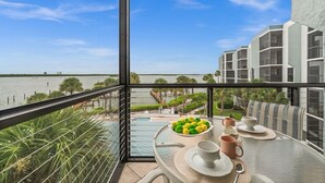 A balcony with a table set for breakfast, overlooking a body of water and a modern building. - Bayfront Condo w/ Sunrise Views & Resort Amenities (Marco Island)