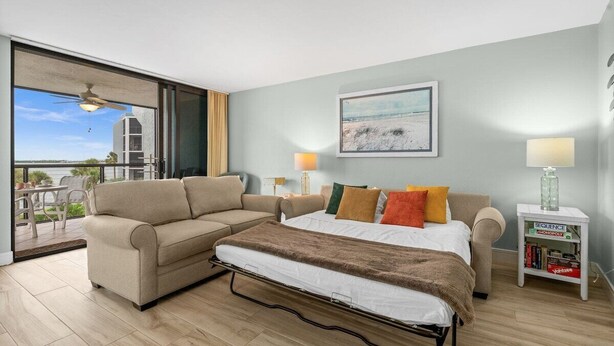 Une chambre à coucher avec un lit, un canapé, une table de chevet avec des livres et une vue à travers une porte vitrée.