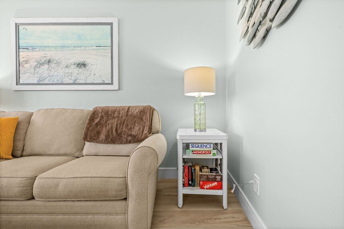 Un salon avec un canapé beige, une petite table d’appoint blanche sur laquelle se trouvent une lampe et des jeux de société, et un tableau encadré accroché au mur.